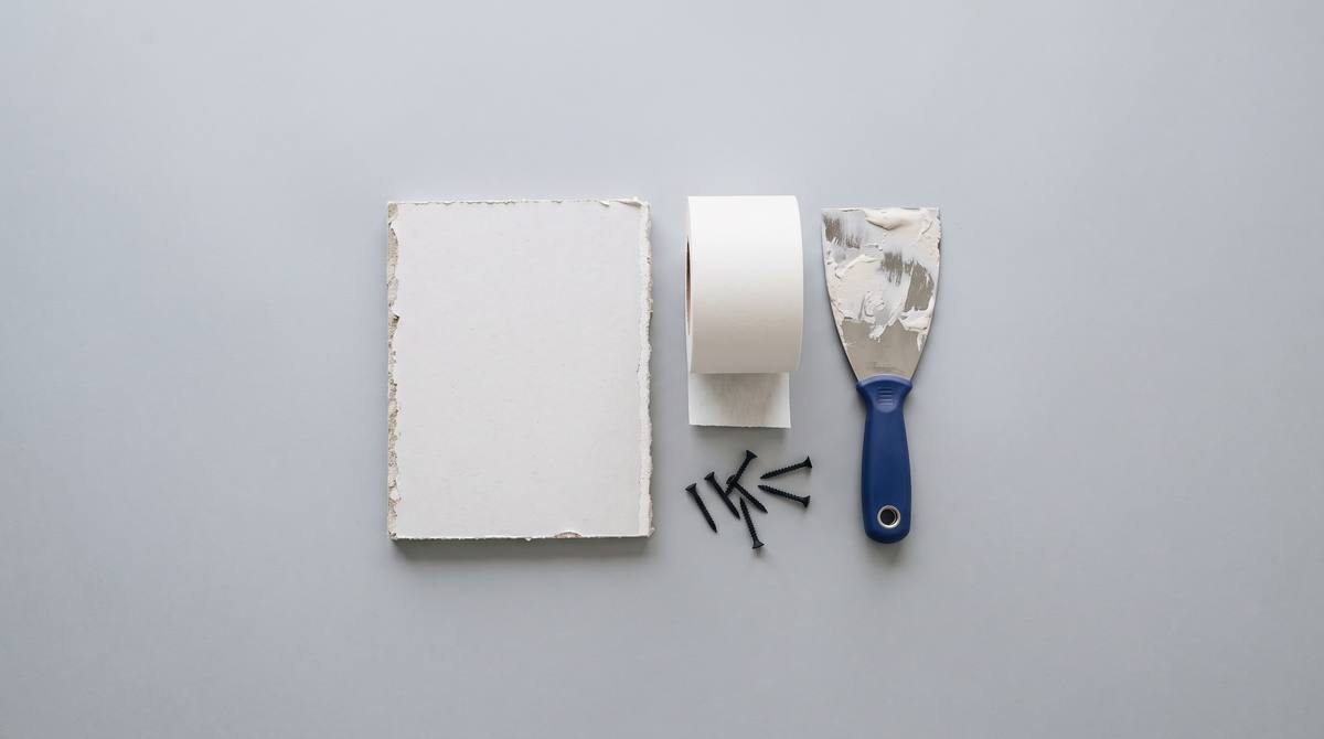 A piece of drywall, a roll of joint tape, a taping knife and drywall screws on grey paper