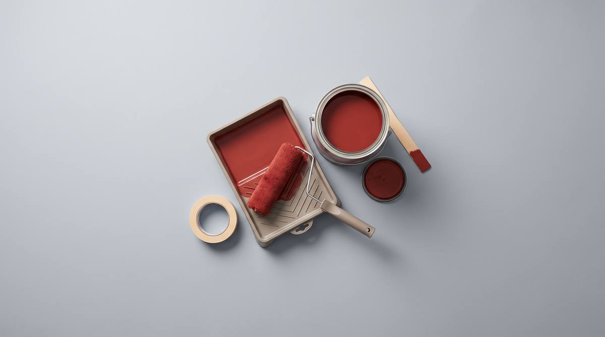 An open can of red paint, a roller in a paint tray, painter’s tape and a stir stick on grey paper